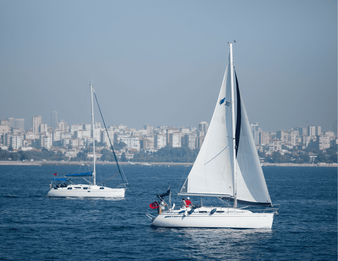 Dos veleros blancos navegando en aguas azules con el horizonte de una ciudad costera llena de edificios en el fondo